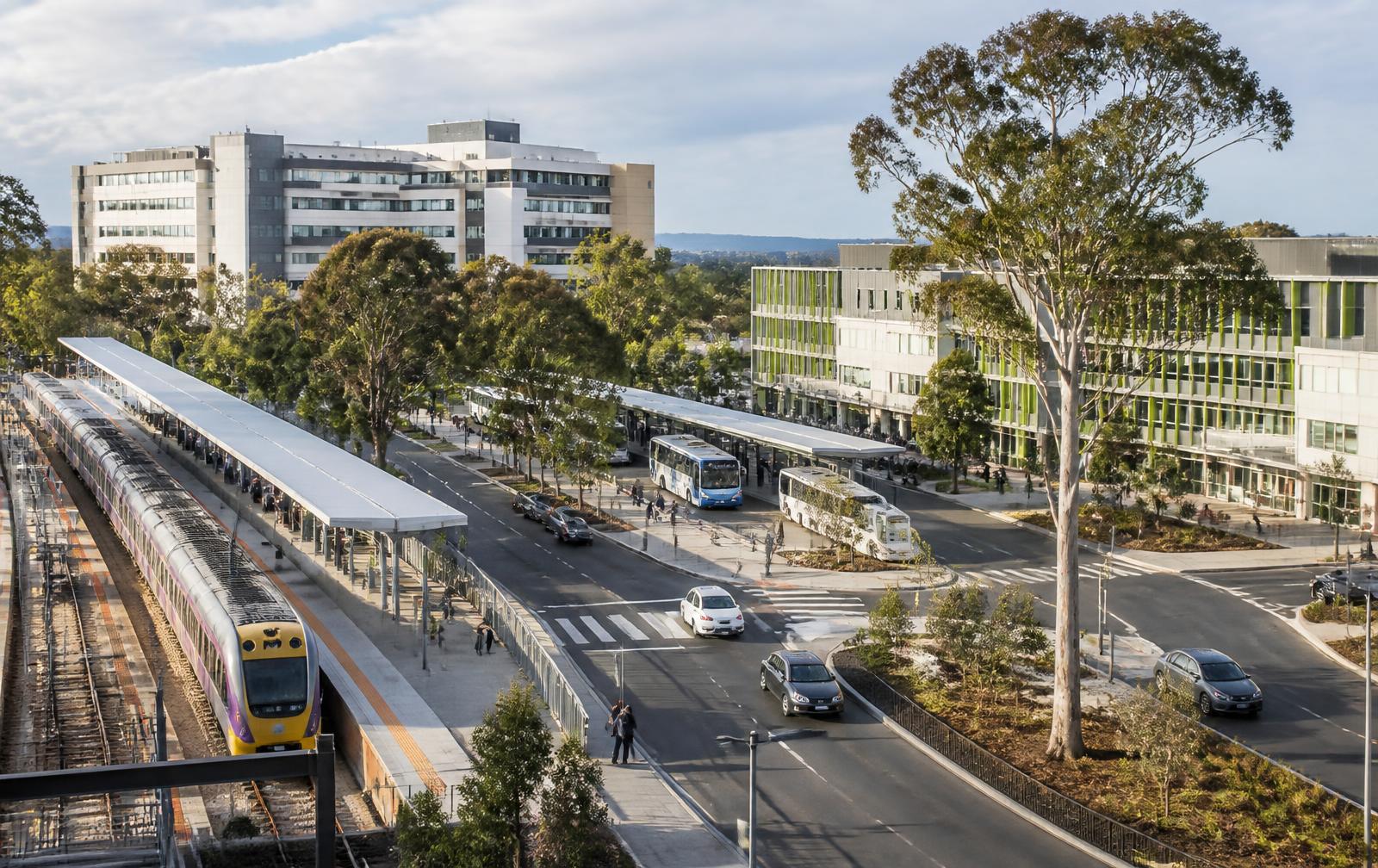 Regional Australian transport interchange with a train at the platform, buses, and adjacent hospital and education buildings