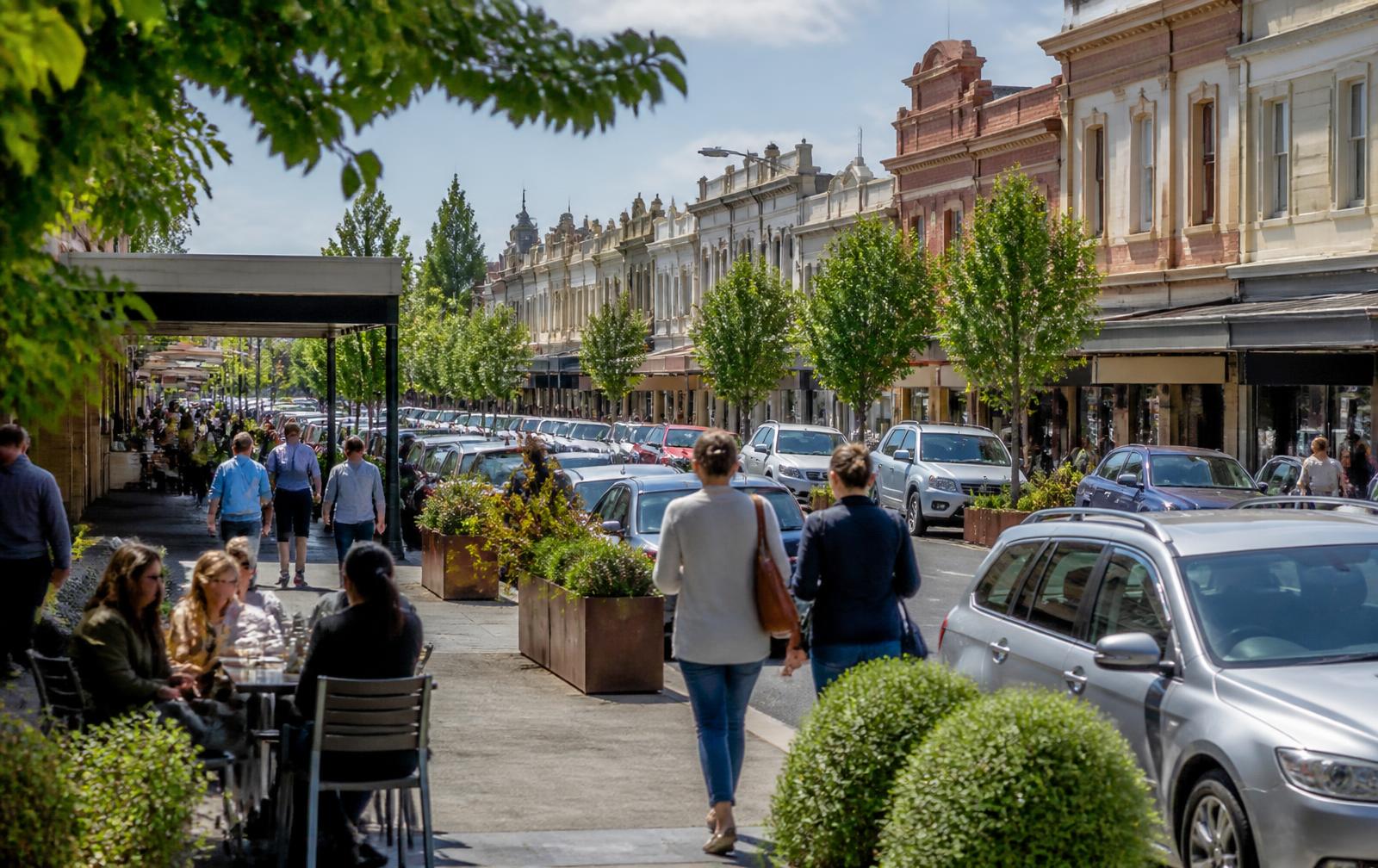 Busy Australian regional town centre street with shopfronts, cafes, street trees and pedestrians